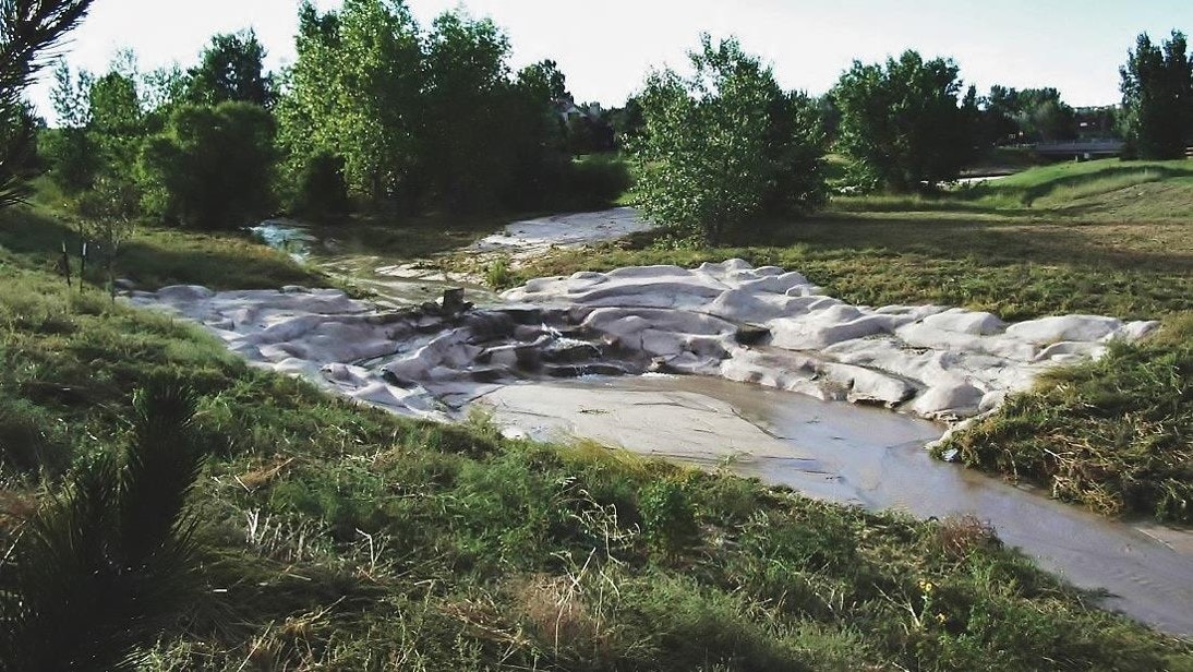 A stream flowing through molded concrete beds in a landscaped area with trees and vegetation.