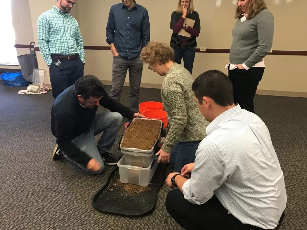 Group of people participating in an indoor activity, possibly a team-building exercise involving containers of soil and rocks.