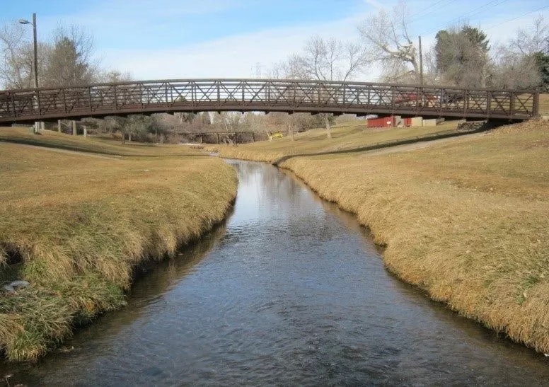 A metal bridge over a narrow river with grassy banks and trees in the background.