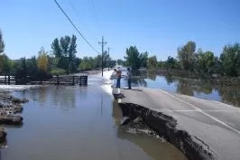 A flooded road with a collapsed section and two individuals observing the damage.