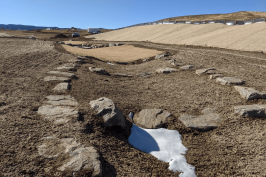 A barren landscape with patches of snow, rocks, and distant buildings under a clear blue sky.