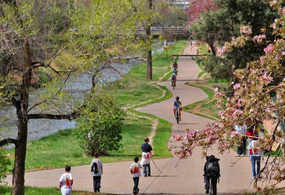 People walking and cycling on a park path by a river, with blooming trees.