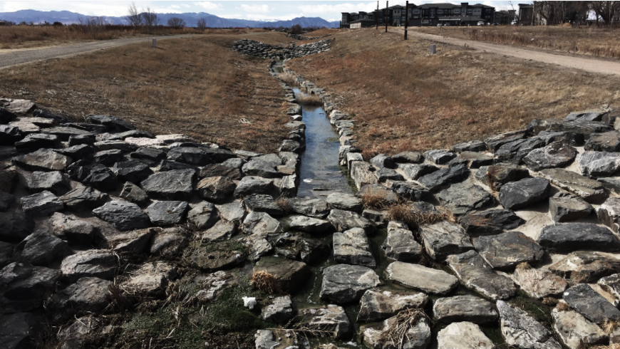 A stone-lined channel with a trickle of water, grassy banks, and a backdrop of cloudy skies.