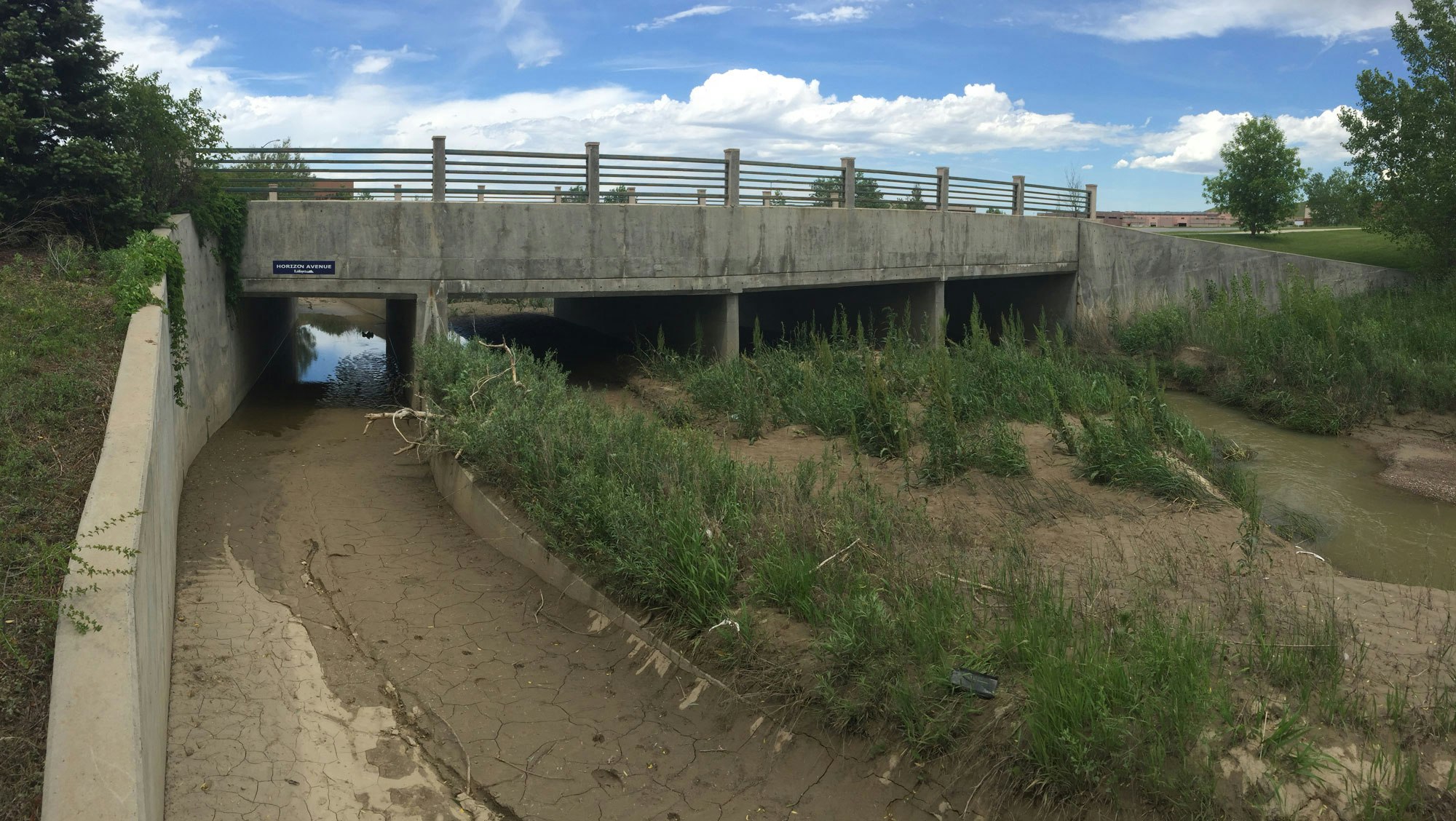 A concrete bridge over a small waterway with surrounding greenery and a dried-up area with cracked soil.