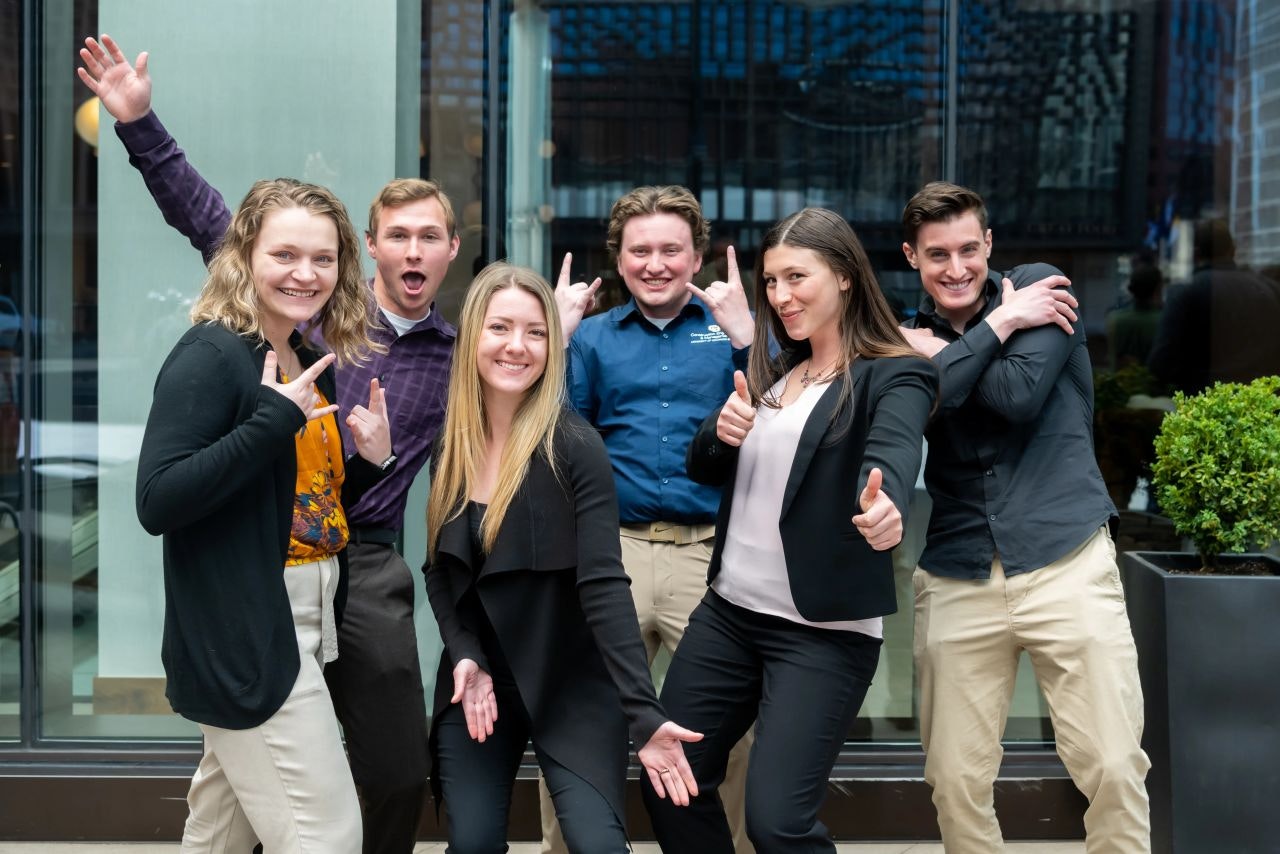 A group of six people posing playfully, smiling and giving thumbs up in front of a glass building.
