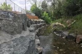 A stream flowing over rocks with greenery and a fence in the background.