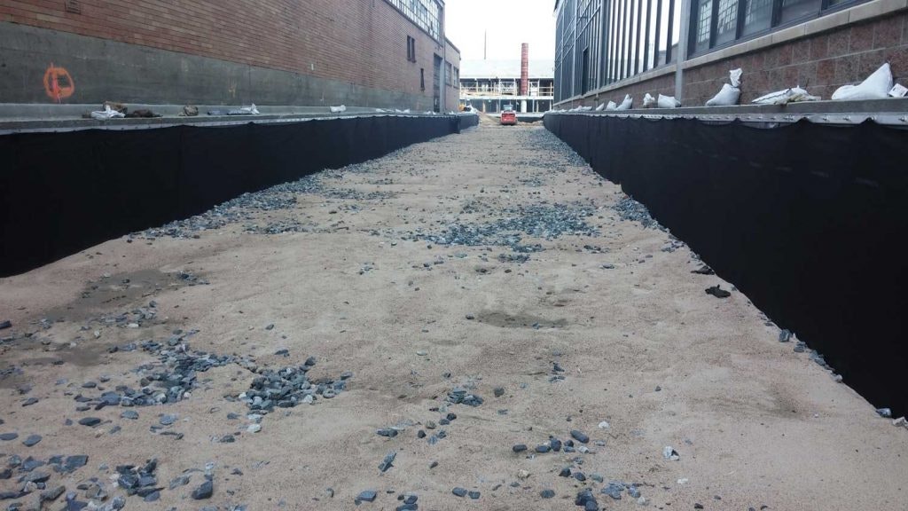 A construction site with sand, scattered stones, and a covered trench between buildings.
