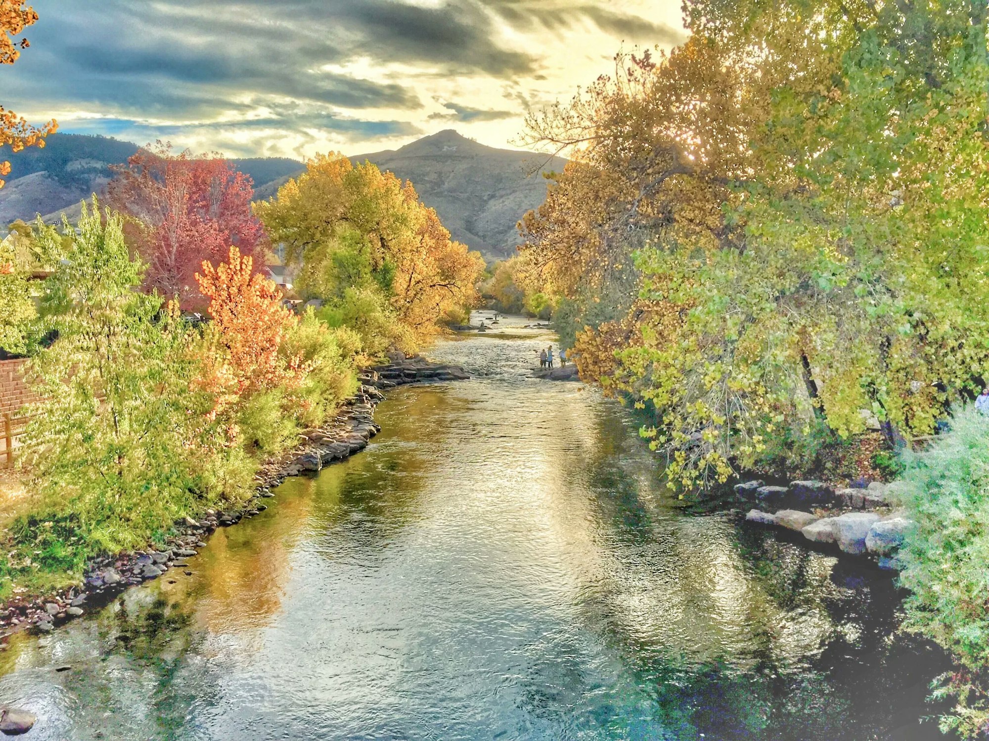 Autumnal trees line a serene river with mountains in the background and a cloudy sky above.