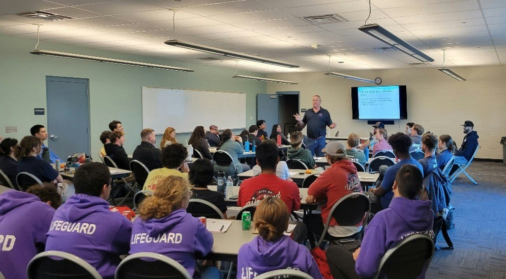 A group of lifeguards is attending a training session, listening to a speaker in a classroom setting.