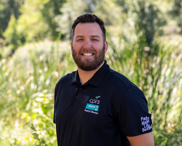 A person with a beard in a black shirt stands outdoors with greenery in the background.