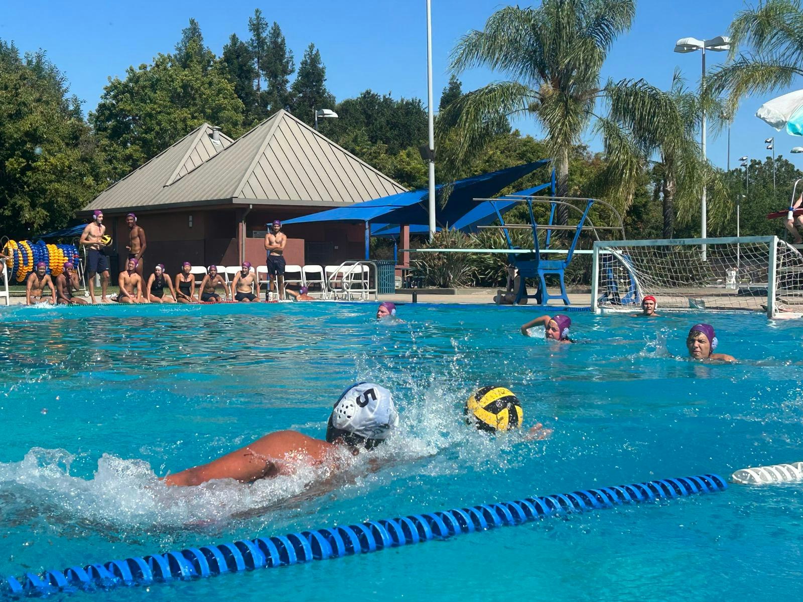 A water polo match in progress with players in caps and spectators by the pool.