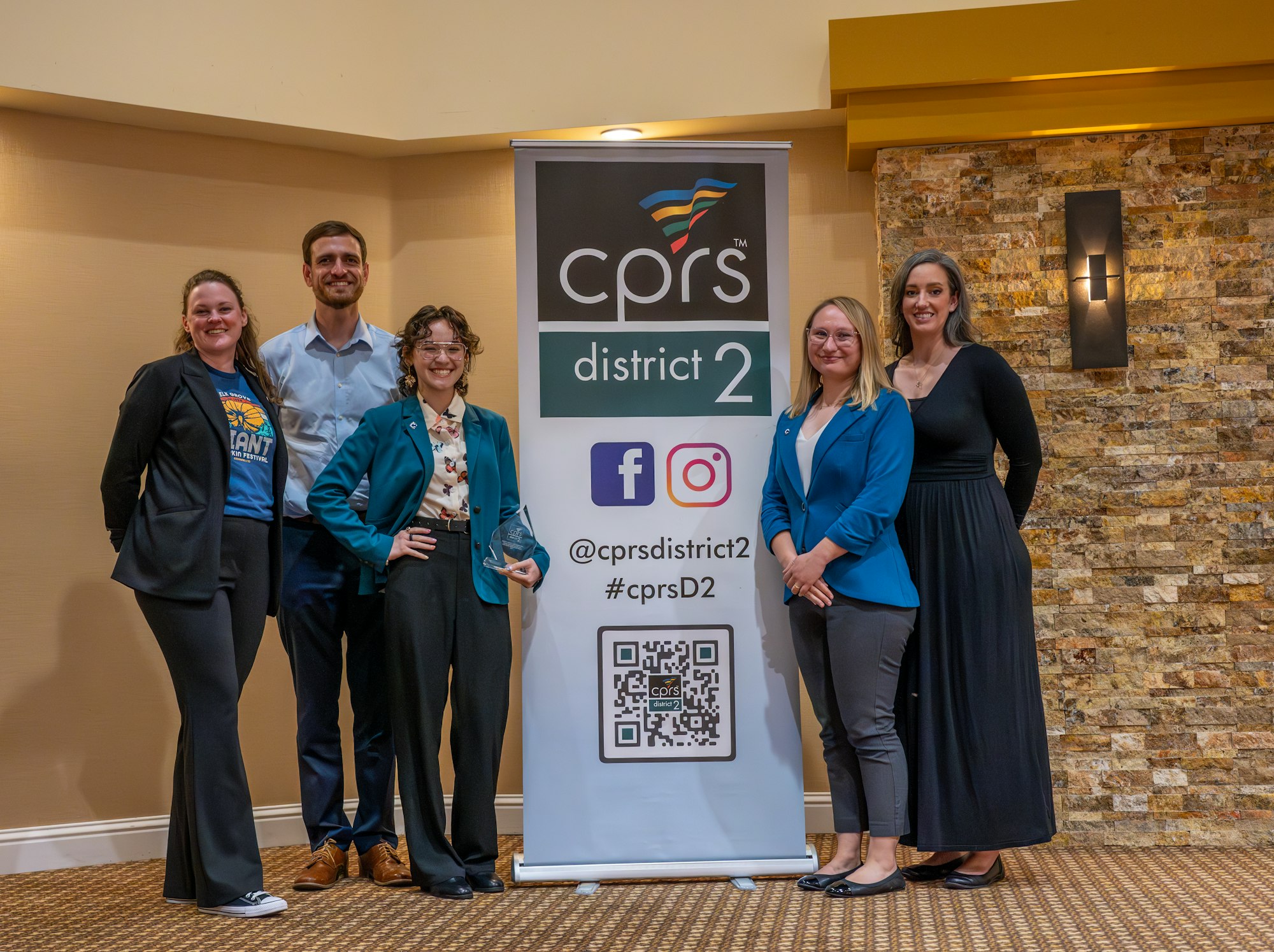 A group of five people stands next to a banner for CPRS District 2, highlighting social media and contact info.
