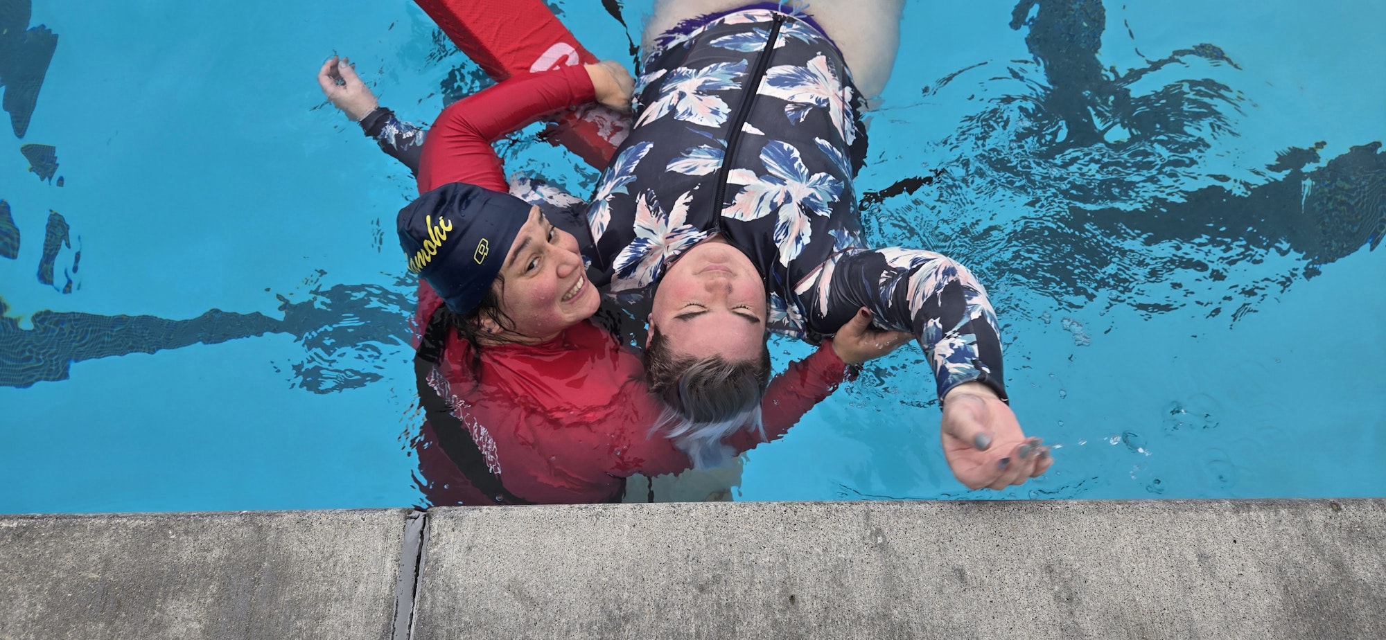 Two people in a pool practicing a rescue maneuver, one holding the other afloat near the pool edge.