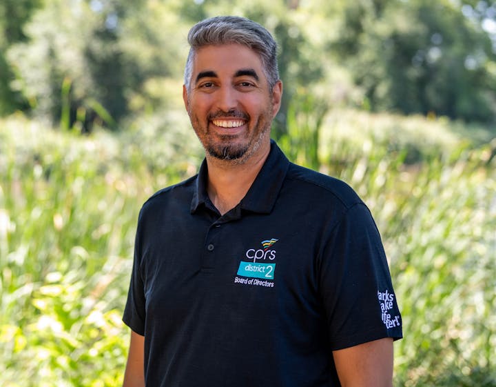 A person smiling outdoors wearing a black shirt with CPRS District 2 and Board of Directors logos.