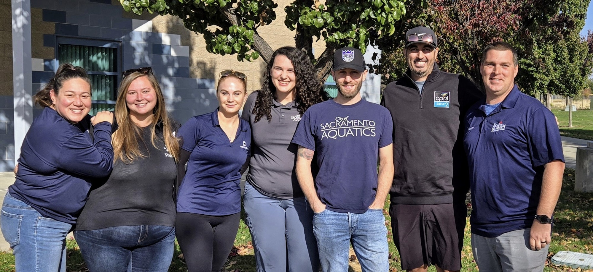 Seven people posing for a photo outdoors, some wearing shirts with "Sacramento Aquatics" logos.