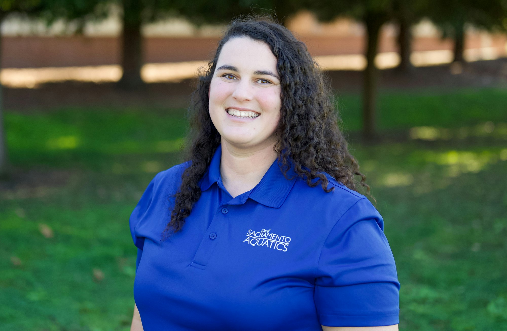 A person in a blue shirt with "Sacramento Aquatics" logo, smiling outdoors with trees in the background.
