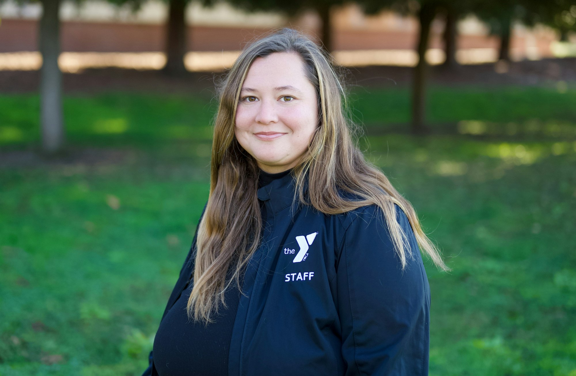 A person smiling, wearing a jacket with "The Y STAFF" on it, standing outdoors with grass and trees in the background.