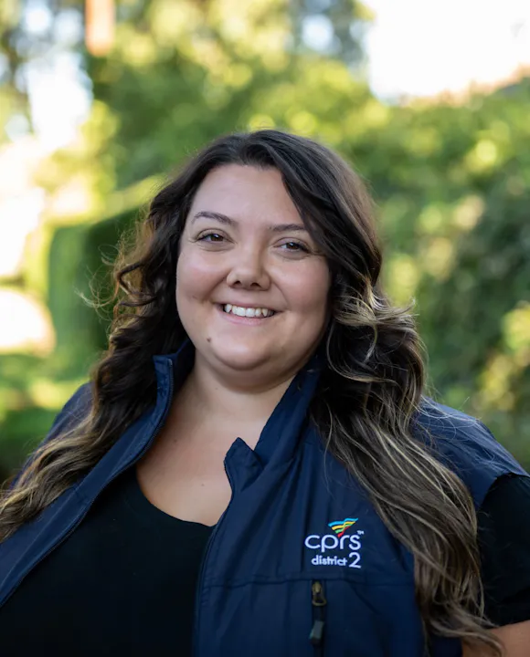 A smiling woman in a blue vest with the logo "cprs district 2" stands outdoors against a blurred green background.