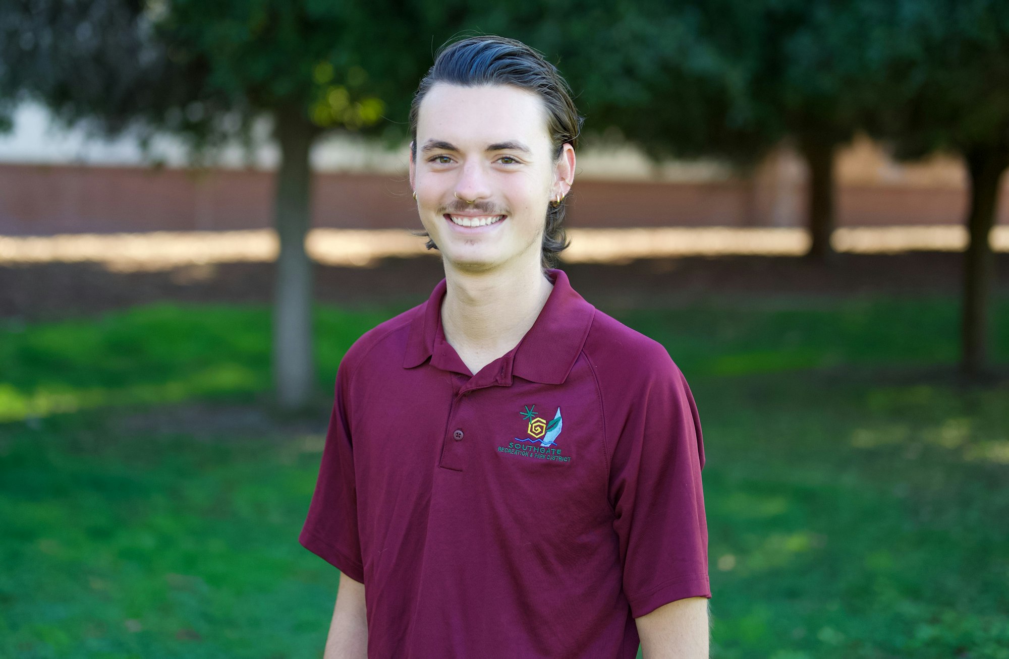 A person smiling outdoors, wearing a maroon polo shirt with a logo.