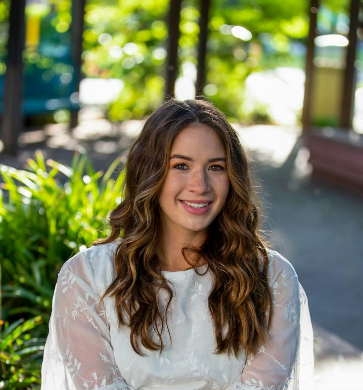 Smiling person with long hair wearing a white top, sitting outdoors with greenery and sunlight in the background.