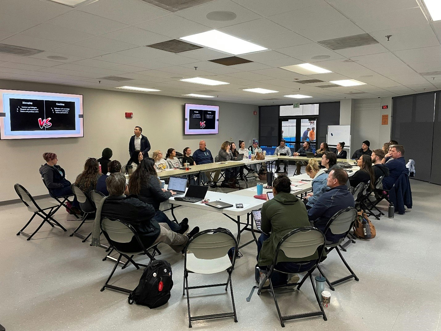 A group of people in a meeting, seated in a U-shape around tables, with a presentation displayed on screens.