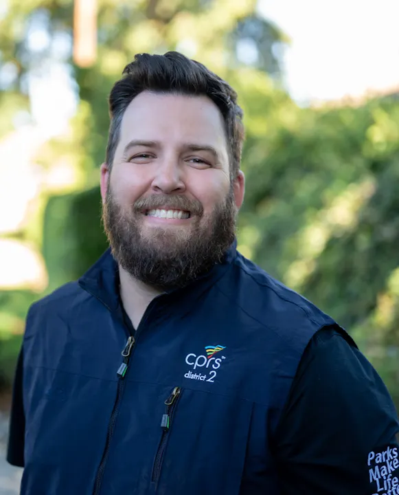A smiling man with a beard, wearing a dark vest with "cprs district 2" logo, stands outdoors with greenery in the background.
