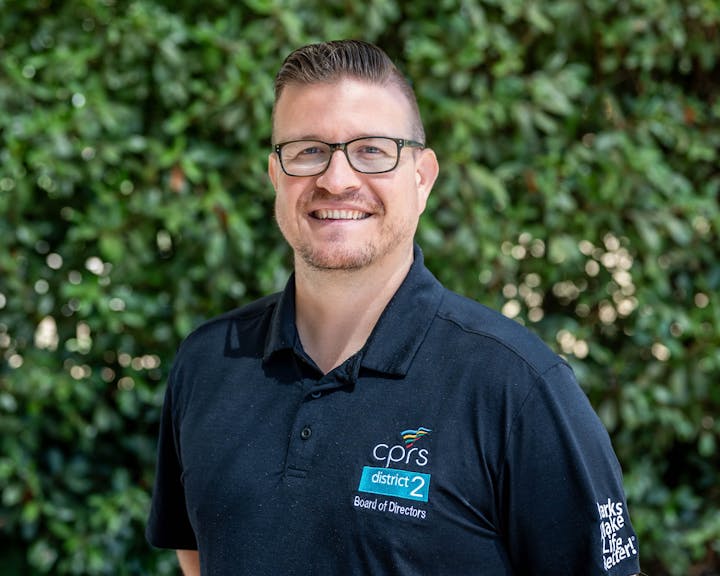 A person smiling, wearing glasses and a CPRS District 2 Board of Directors shirt, with a leafy background.