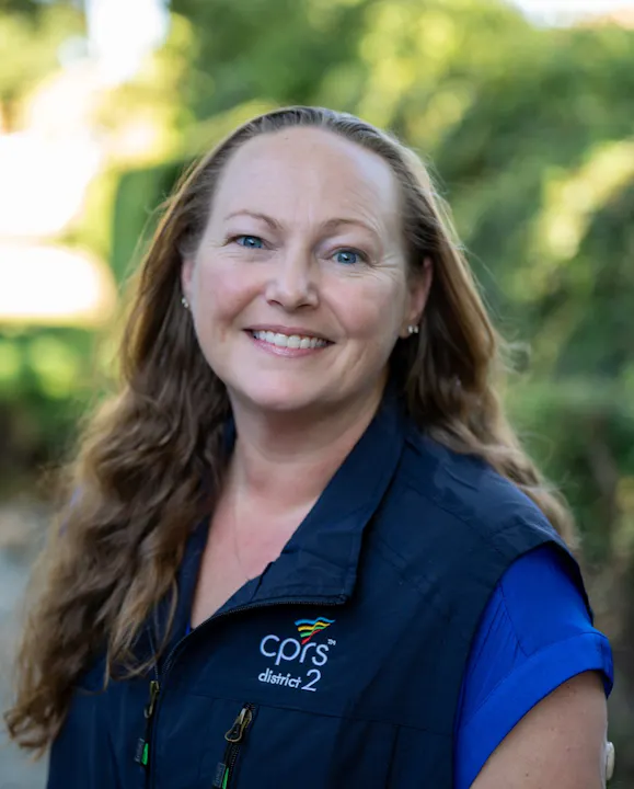 A smiling woman with long, wavy hair is wearing a navy vest with a logo, set against a green background.