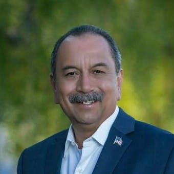 A person in a suit with a flag pin, smiling in an outdoor setting.