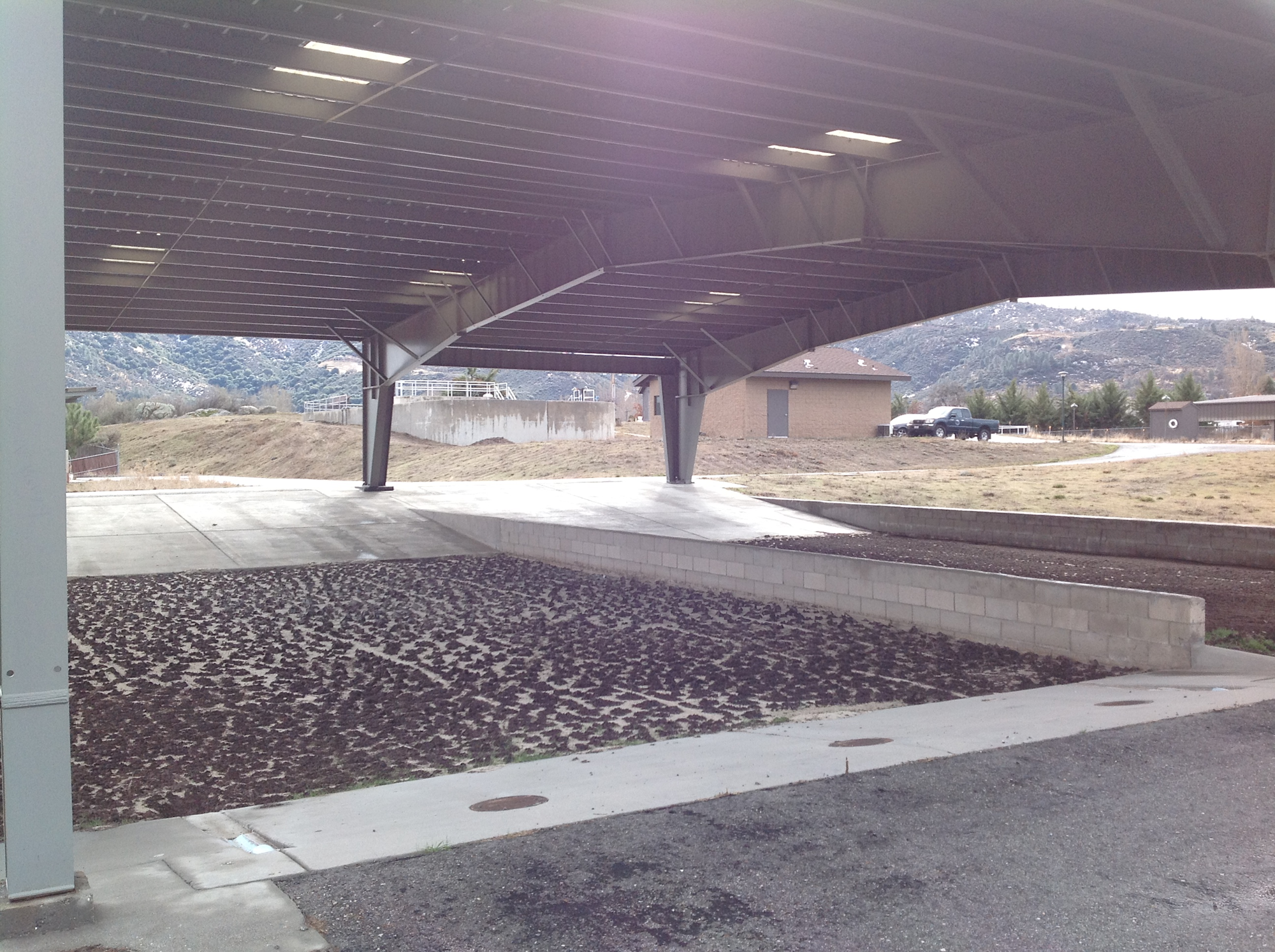 An open structure with a metal roof, concrete ground, and surrounding buildings, set in a rural area with hills in the background.