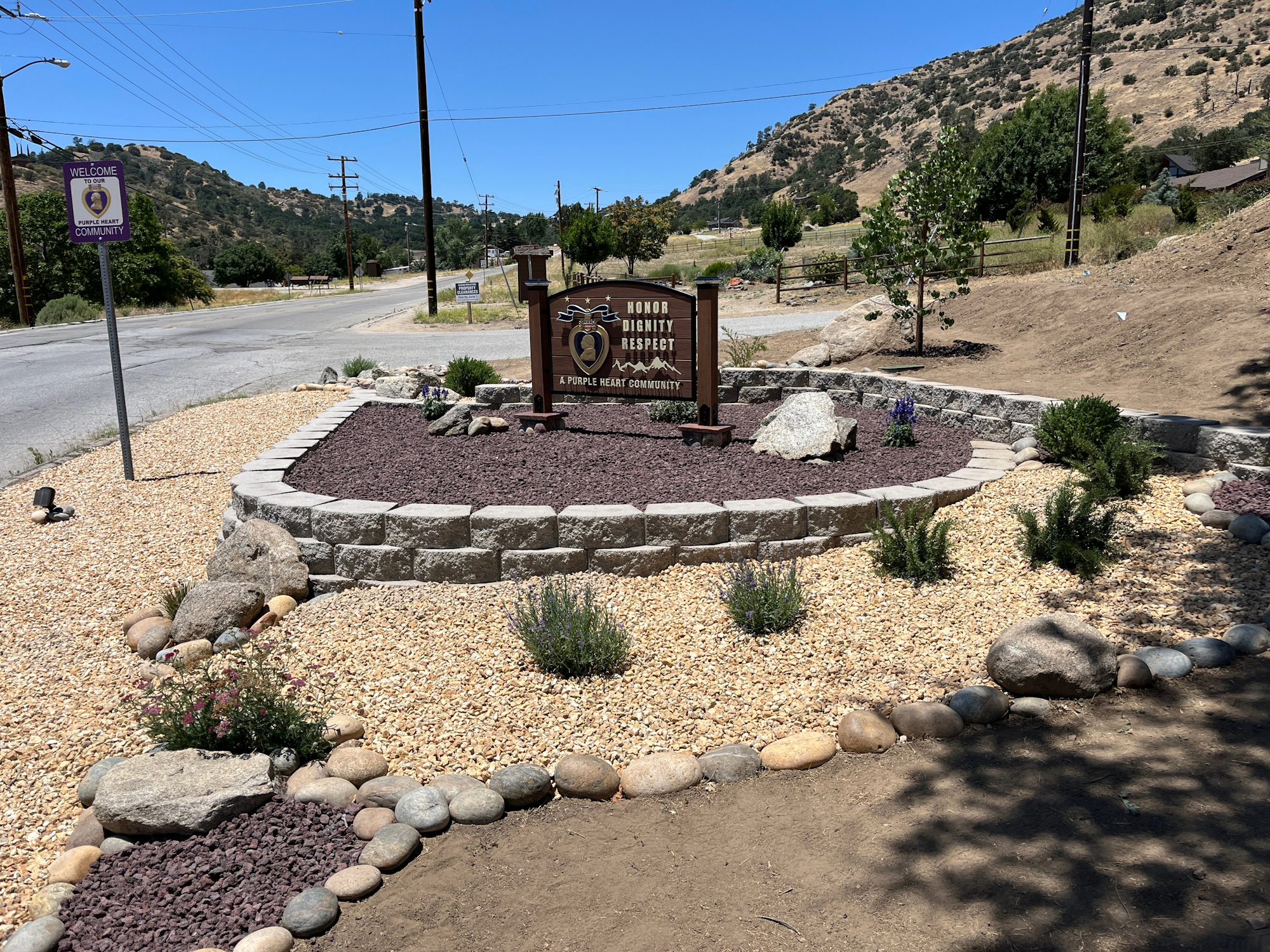 A landscaped area with a sign stating "Honor, Dignity, Respect" and nearby road, trees, and hills under a clear blue sky.