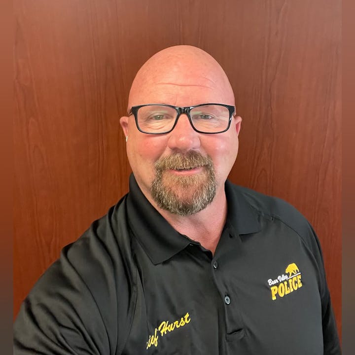 A person with glasses, wearing a black "Bear Valley Police" shirt, standing in front of a wooden wall.