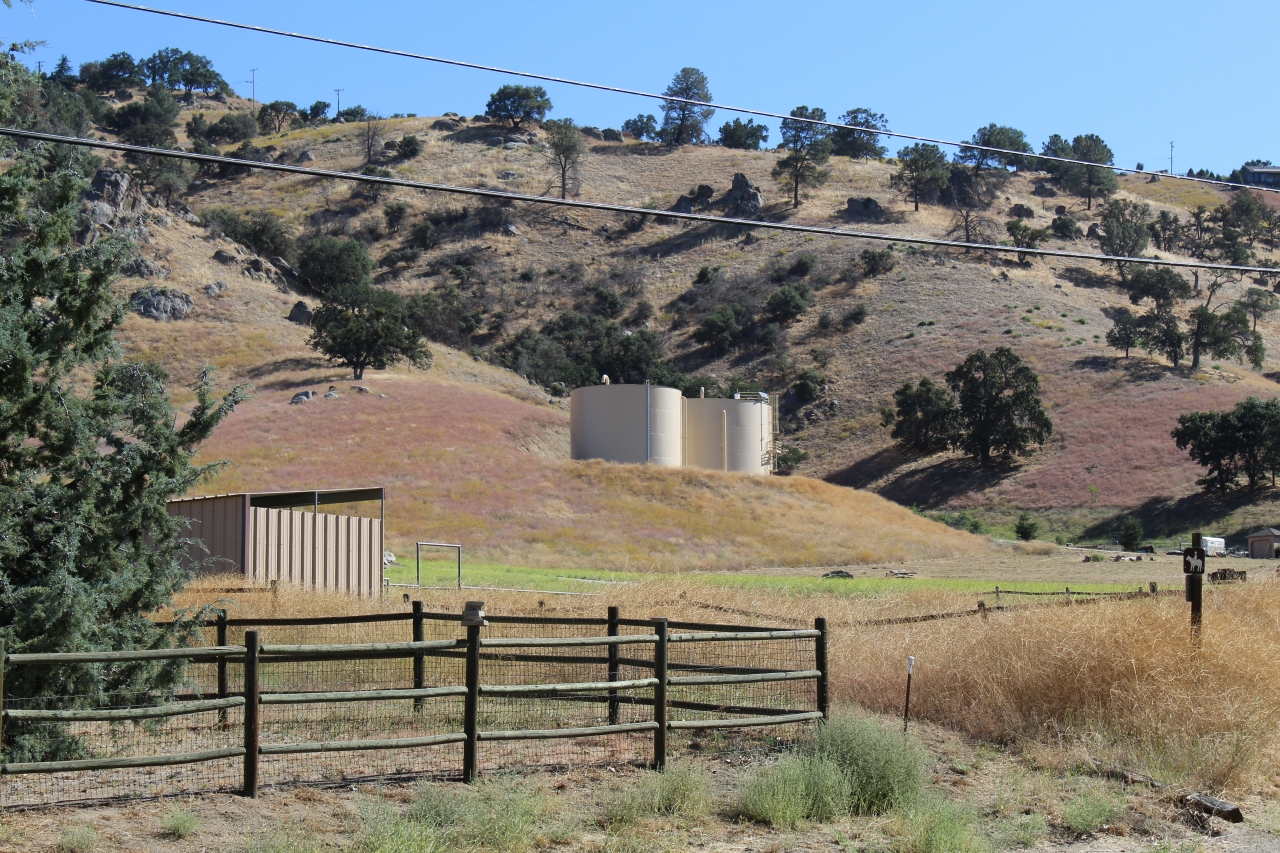 Hilly landscape with trees, a fenced area, utility structures, and storage tanks in the background.