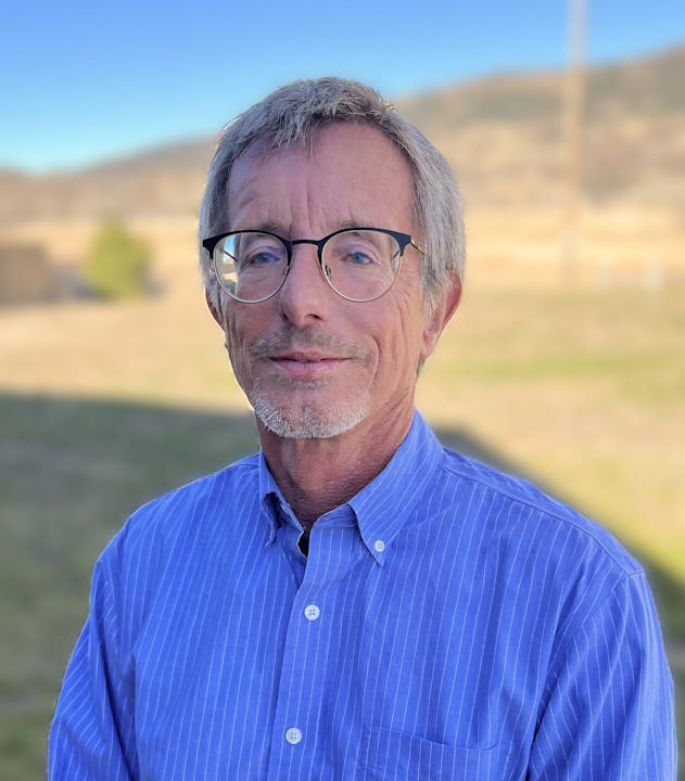 A person wearing glasses and a blue shirt standing outside with a blurred natural background.