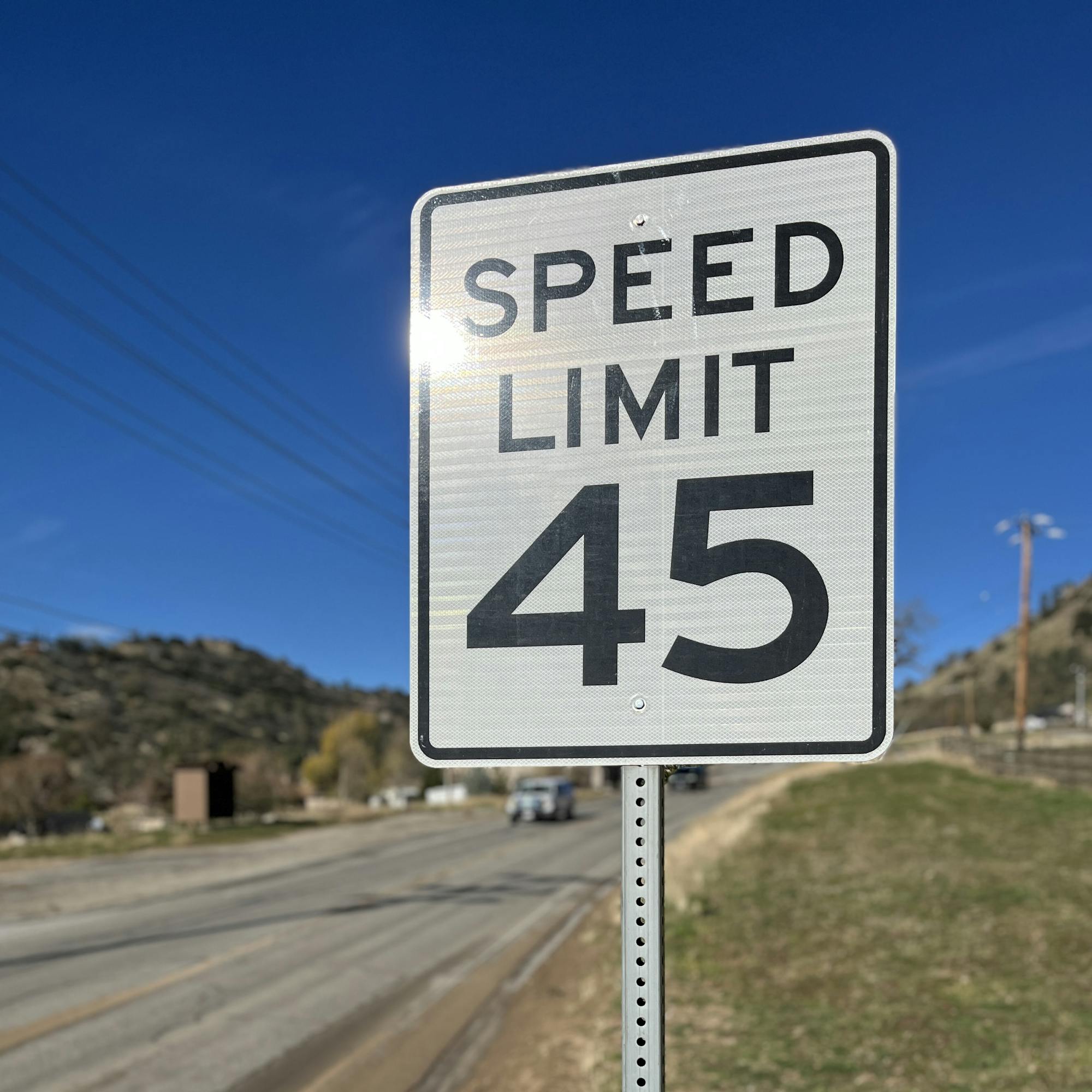 A speed limit sign indicating a maximum speed of 45 mph along a road, with hills and a clear blue sky in the background.