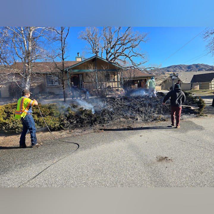 People are working to extinguish smoldering debris near a house, with smoke rising against a clear blue sky.