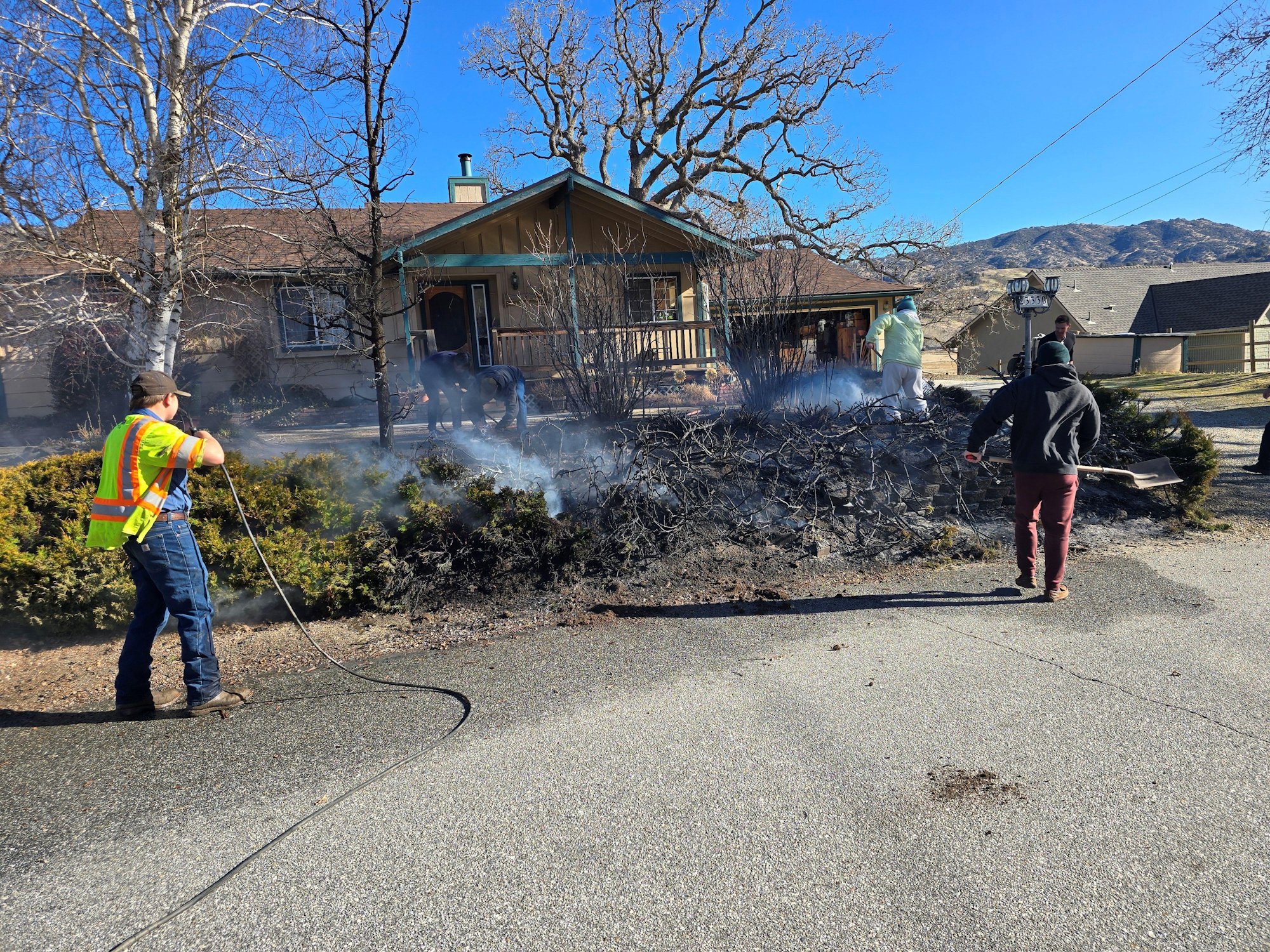 People are working to extinguish smoldering debris near a house, with smoke rising against a clear blue sky.