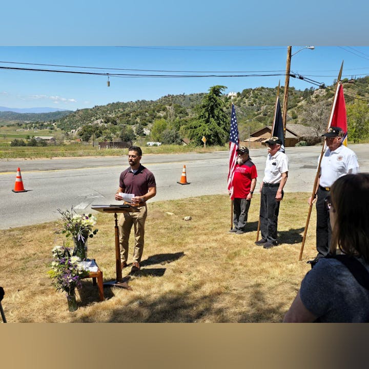 A person speaks at podium outdoors, three people hold flags; there are flowers and audience, with hills in the background.