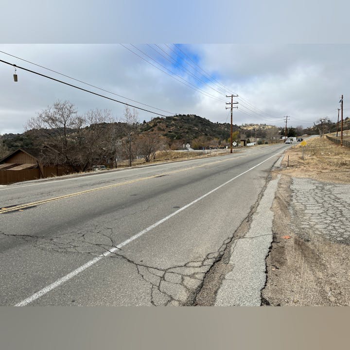 A cracked, empty road with power lines, trees, and hills under a cloudy sky.