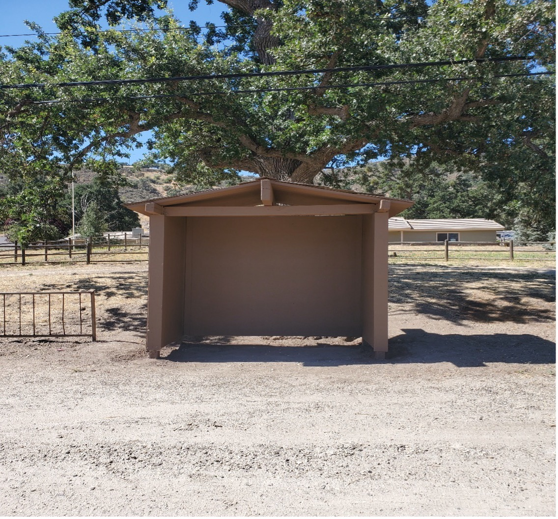 A small brown shelter under a tree by a fence, with a house in the background.