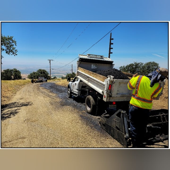 A worker operates a truck spreading gravel or asphalt on a rural road in a scenic area.