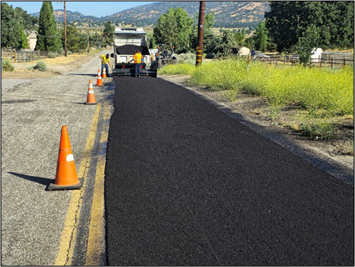 Roadwork with fresh asphalt being laid, traffic cones, workers, and a truck on a rural road.