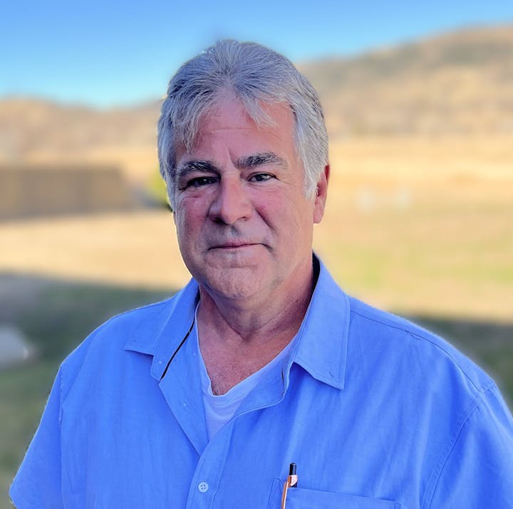 A person with gray hair in a blue shirt stands outdoors with a blurry background of grass and hills.