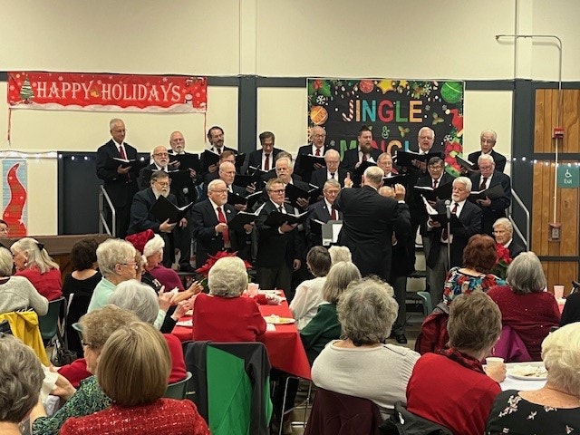 A choir performs for an audience at a holiday gathering, with festive decorations and attendees enjoying refreshments.