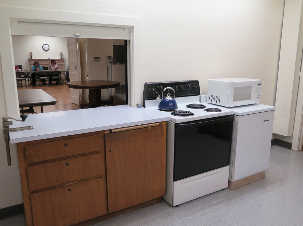A kitchen with a stove, a blue kettle, a microwave, and cabinets. There's an opening to a room with people and tables.