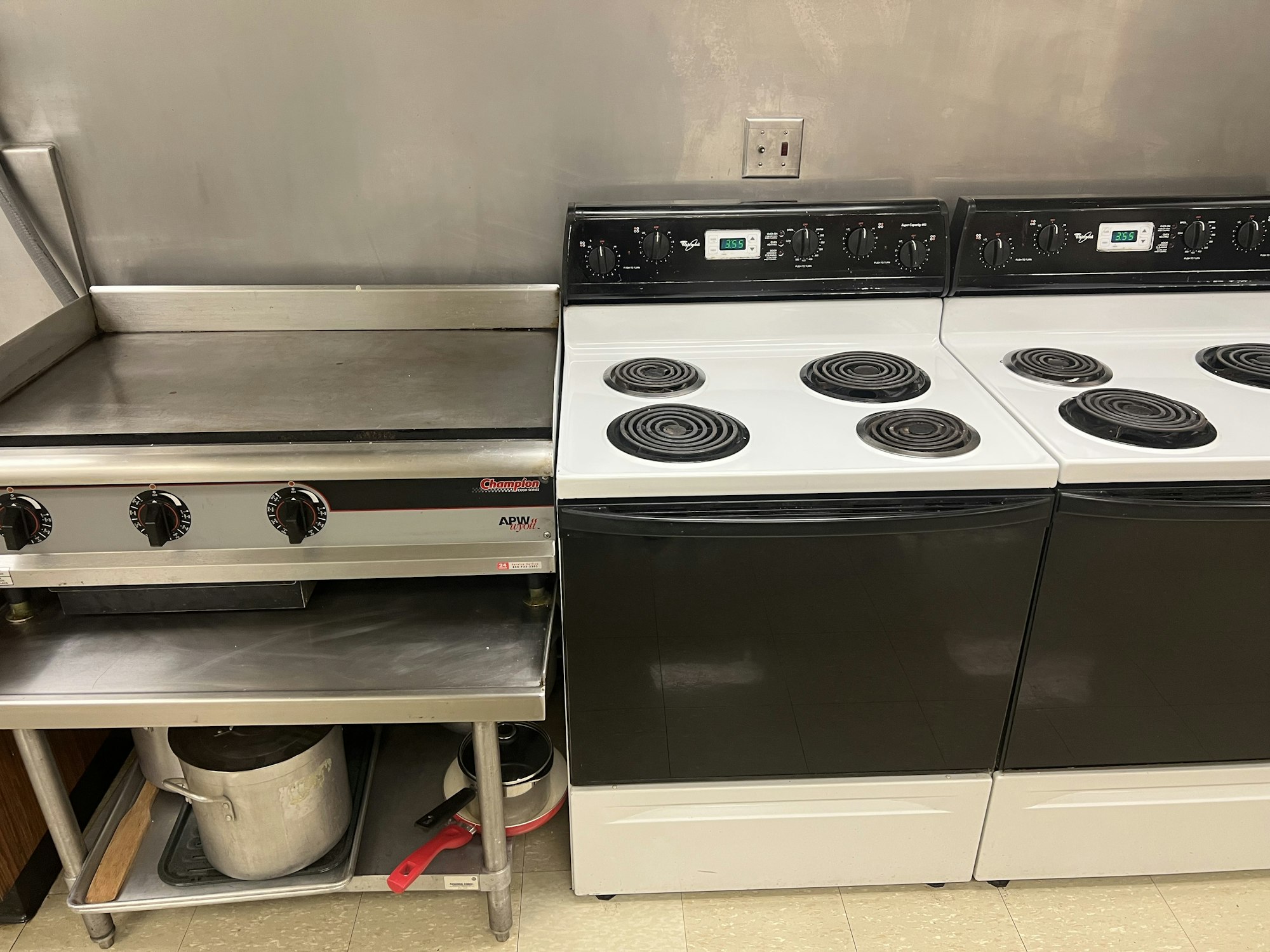 A kitchen with a griddle, two electric stoves, a pot, and some cooking utensils.
