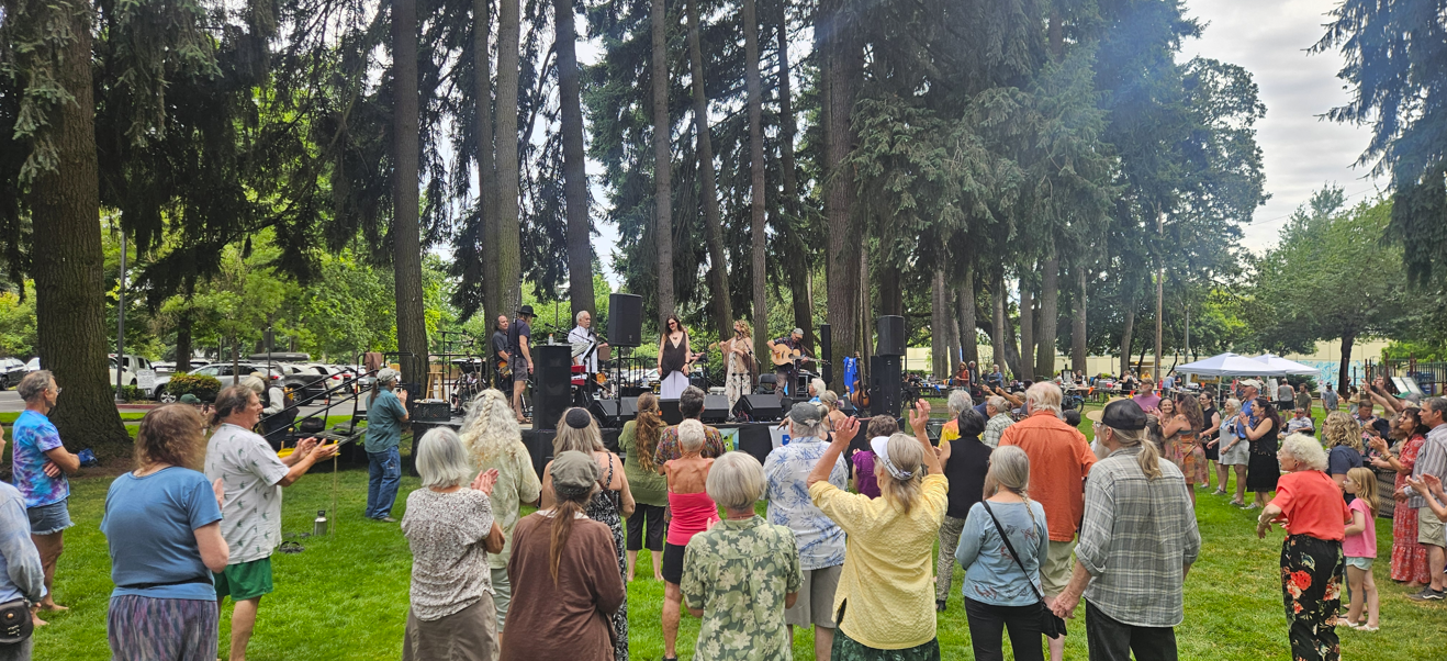 A lively outdoor music event with a performance on stage and an engaged audience enjoying the atmosphere among tall trees.