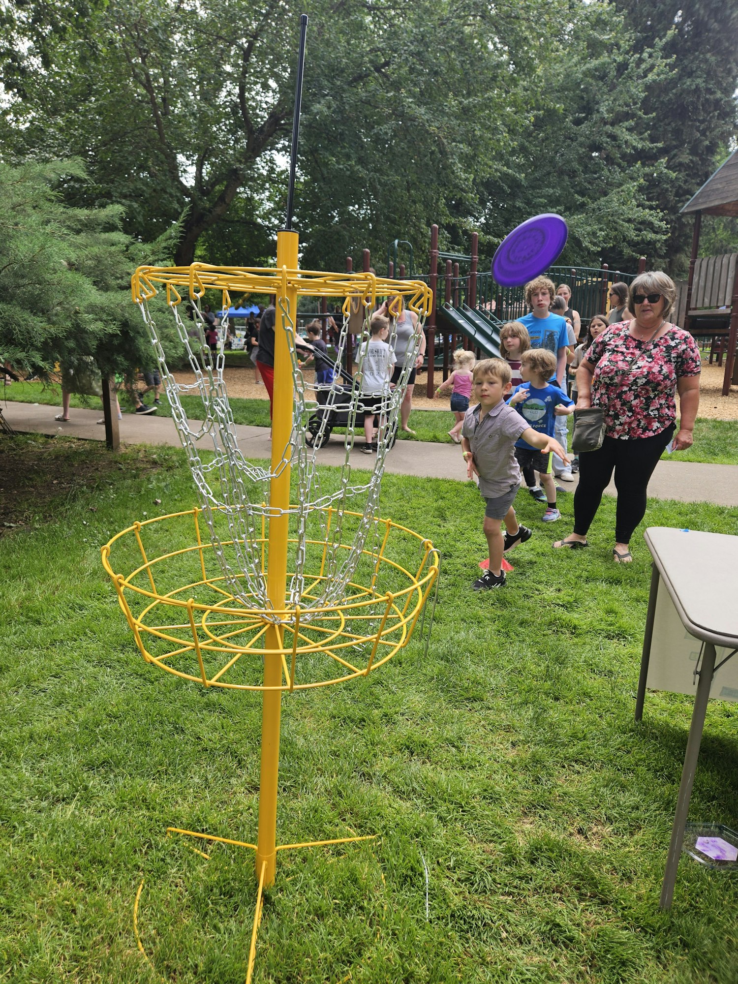 A child throws a purple frisbee towards a yellow disc golf basket in a park with a group of people watching.