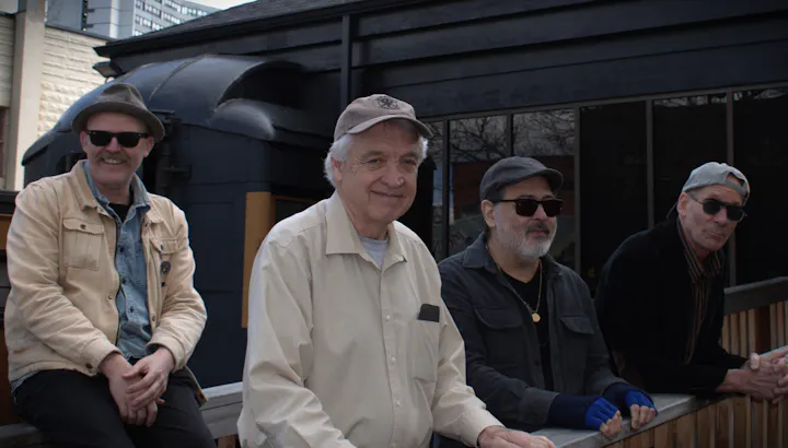 Four men are sitting on a railing, each wearing sunglasses and casual attire, with a dark building in the background.