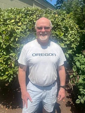 Man smiling, wearing "OREGON" shirt, standing in front of green bushes under sunny sky.